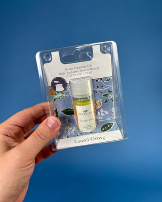 A hand holds a packaged bottle of Laurel Grove home fragrance oil against a solid blue background. The product is in a clear blister pack with a white backing card featuring floral illustrations.