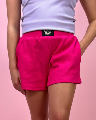 Person wearing bright pink shorts with a visible brand label on a pink background