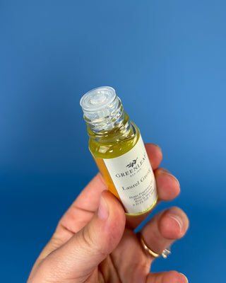 A hand holding a small glass bottle of Greenleaf Organics Laurel Green oil against a solid blue background.