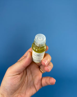 A hand holding a small glass bottle filled with yellow-green liquid against a solid blue background. The bottle has a white label and a clear cap.