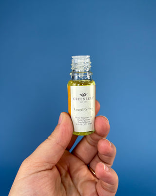 A hand holding a small glass bottle of Greenleaf Organics Laurel Green oil against a solid blue background.
