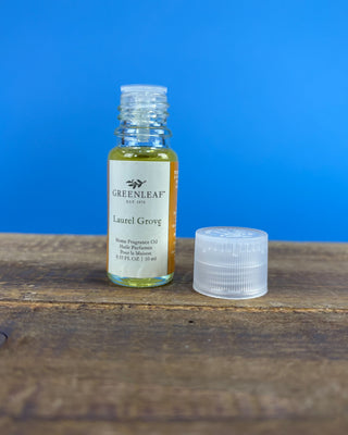 A small clear bottle of Greenleaf Laurel Grove home fragrance oil with a cream label, standing on a rustic wooden surface next to its clear ribbed cap against a solid blue background.