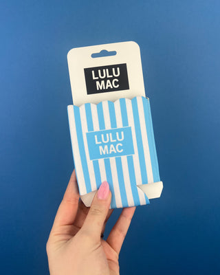 A hand with pink nail polish holds a Lulu Mac branded sleeve or holder against a solid blue background.