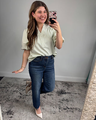 Woman taking a mirror selfie wearing a striped shirt and jeans in a room with a gray wall.