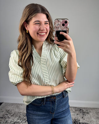 Woman taking a selfie in a striped shirt and jeans against a gray wall.