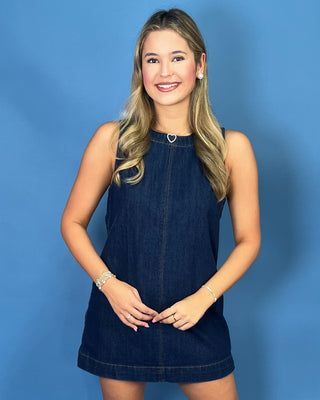 A smiling young woman with blonde hair models a sleeveless dark blue denim mini dress 