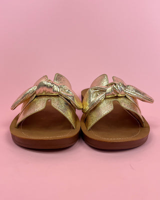 Pair of gold sandals with bow details on a pink background