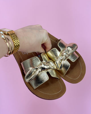 Gold sandals with bows held by a hand against a pink background
