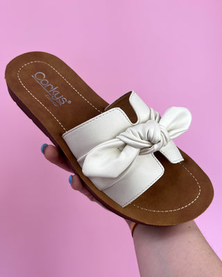 Brown sandal with a white bow held against a pink background