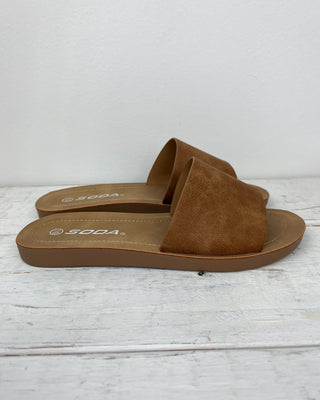 Brown sandals with 'Soda' branding on a light wooden floor.