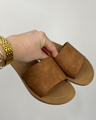 Brown sandal held by a hand with gold bracelets on a light gray background