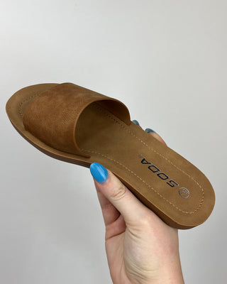 Brown slide sandal held by a hand with blue nail polish on a light gray background