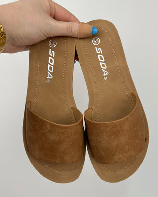 Pair of brown sandals with 'Soda' branding held by a hand on a light gray background
