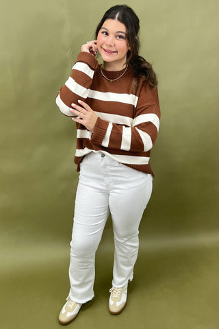 Woman wearing a brown and white striped sweater with white pants against a green background