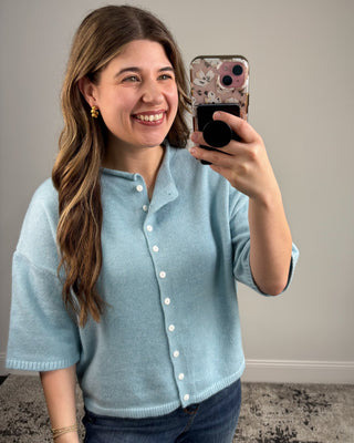 Woman taking a selfie wearing a light blue cardigan against a plain background