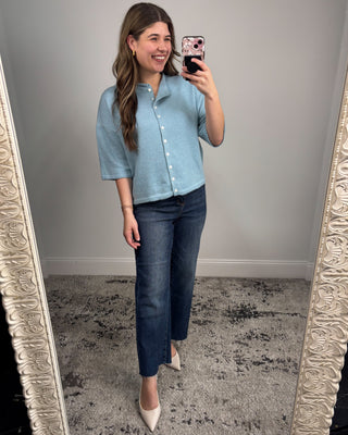 Woman taking a mirror selfie wearing a light blue shirt and dark jeans.