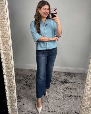 Woman taking a mirror selfie wearing a light blue shirt and dark blue jeans.