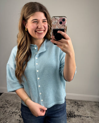 Woman taking a mirror selfie wearing a light blue button-up shirt and jeans.