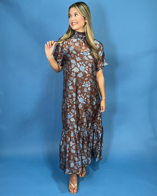 A woman modeling a brown and light blue floral print maxi dress with short flutter sleeves and a tiered ruffle hem