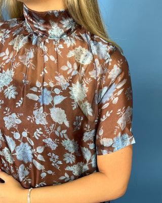 A close-up of a woman wearing a brown short-sleeve blouse with a light blue and white floral print