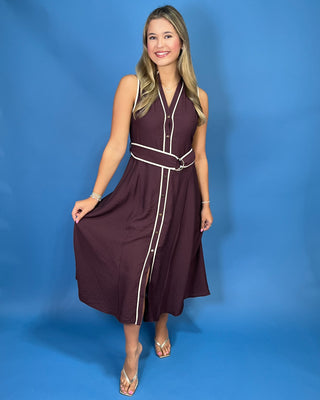 A young woman with blonde hair is wearing a sleeveless dark plum button-up dress with white contrast piping and a matching waist belt