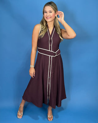 A young woman with blonde hair is wearing a sleeveless dark plum button-up dress with white contrast piping and a matching waist belt