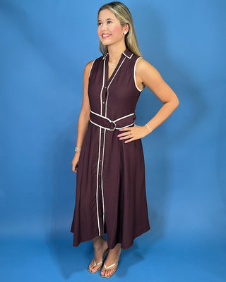 A young woman with blonde hair is wearing a sleeveless dark plum button-up dress with white contrast piping and a matching waist belt