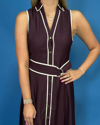 A woman wearing a sleeveless dark plum button-front dress with cream piping trim and a matching waist belt