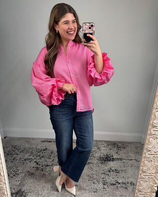Woman taking a mirror selfie wearing a pink blouse with ruffled sleeves and blue jeans.