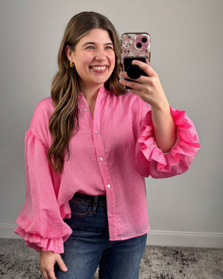 Woman taking a mirror selfie wearing a pink ruffled blouse and blue jeans.