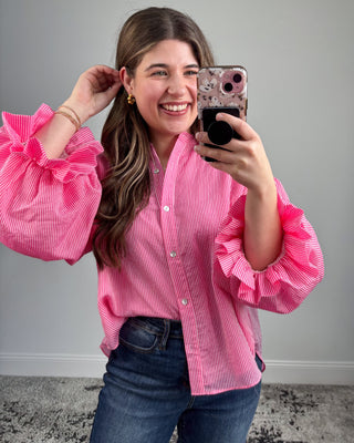 Woman wearing a pink ruffled blouse taking a selfie against a gray wall.