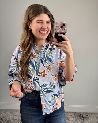 Woman taking a mirror selfie wearing a floral blouse against a plain wall.