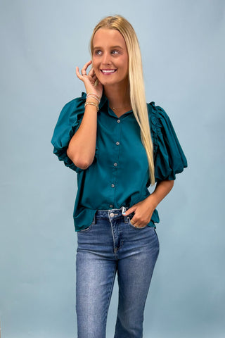 Woman wearing a teal blouse with puffed sleeves and blue jeans against a light gray background
