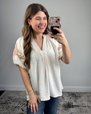 Woman taking a mirror selfie wearing a white blouse with lace sleeves and blue jeans.