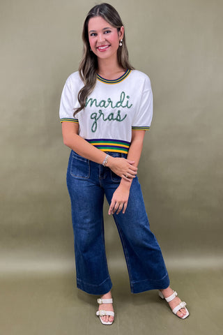 Woman wearing a 'Mardi Gras' shirt and blue jeans against a beige background