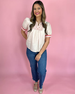 Woman wearing a white blouse with red embroidery and blue jeans on a pink background