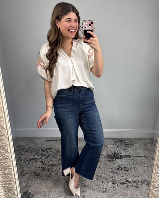 Woman taking a mirror selfie wearing a white blouse and blue jeans in a room with gray walls.