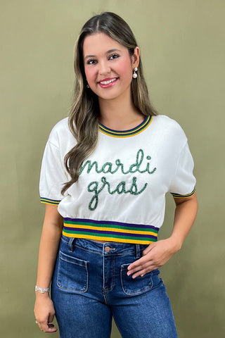 Woman wearing a white t-shirt with 'mardi gras' text and blue jeans against a beige background