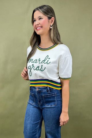 Woman wearing a white sweater with 'Mardi Gras' text and blue jeans against a green background
