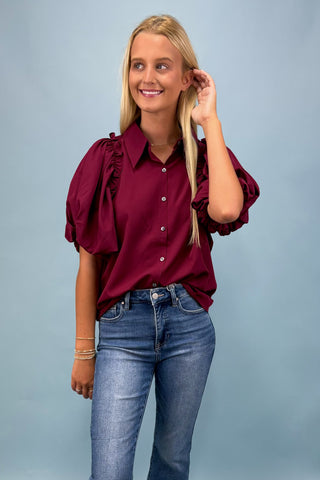 Woman wearing a burgundy blouse with ruffled sleeves and blue jeans on a gray background