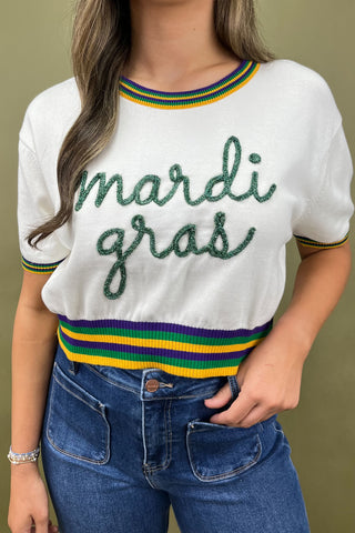 White sweater with 'mardi gras' text and colorful stripes worn by a person against a green background