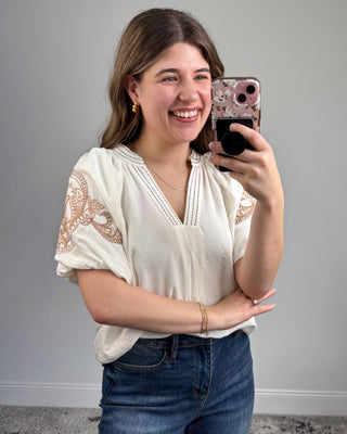 Woman taking a mirror selfie wearing a white blouse with lace sleeves and blue jeans.