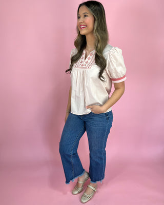 Woman wearing a white blouse with lace details and blue jeans on a pink background