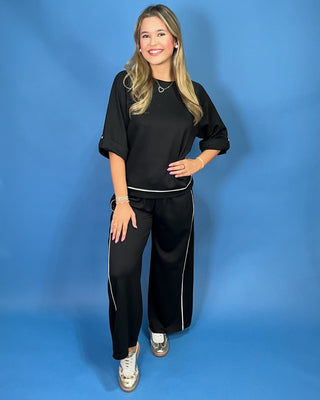 A woman modeling a black two-piece lounge set with white piping details, consisting of a short-sleeve top and matching wide-leg pants