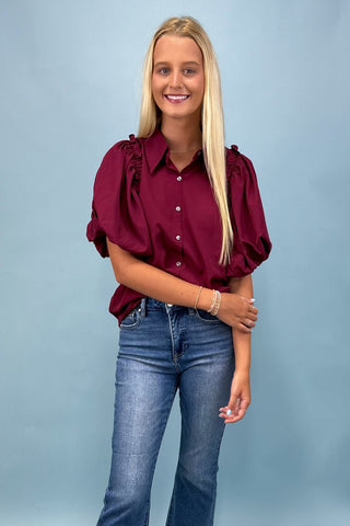 Woman wearing a burgundy blouse with ruffled sleeves and blue jeans against a light blue background