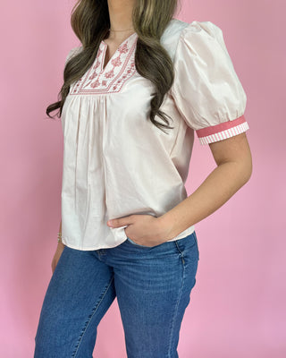 Woman wearing a white blouse with pink embroidery and blue jeans against a pink background