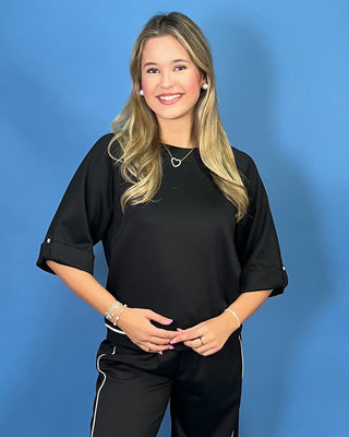 A smiling blonde woman wearing a black short-sleeve top with rolled cuffs and black pants with white side stripes