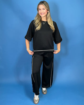 A woman modeling a black two-piece lounge set consisting of a short-sleeve top and matching wide-leg pants with white piping details