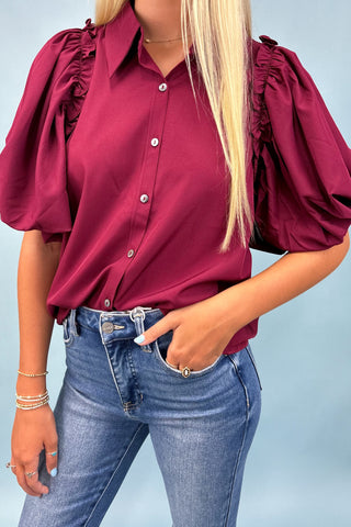 Woman wearing a burgundy blouse with ruffled sleeves and blue jeans against a light blue background