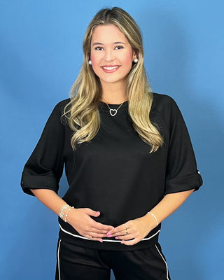 A smiling blonde woman wearing a black short-sleeve top with white piping details and black pants